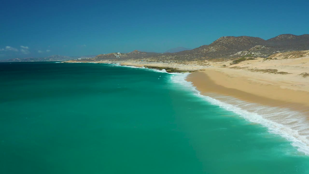 La Laguna Beach in Los Cabos from above, with a broad sandy beach, curling white waves, green-blue water, arid hills across the background, and no visible people.