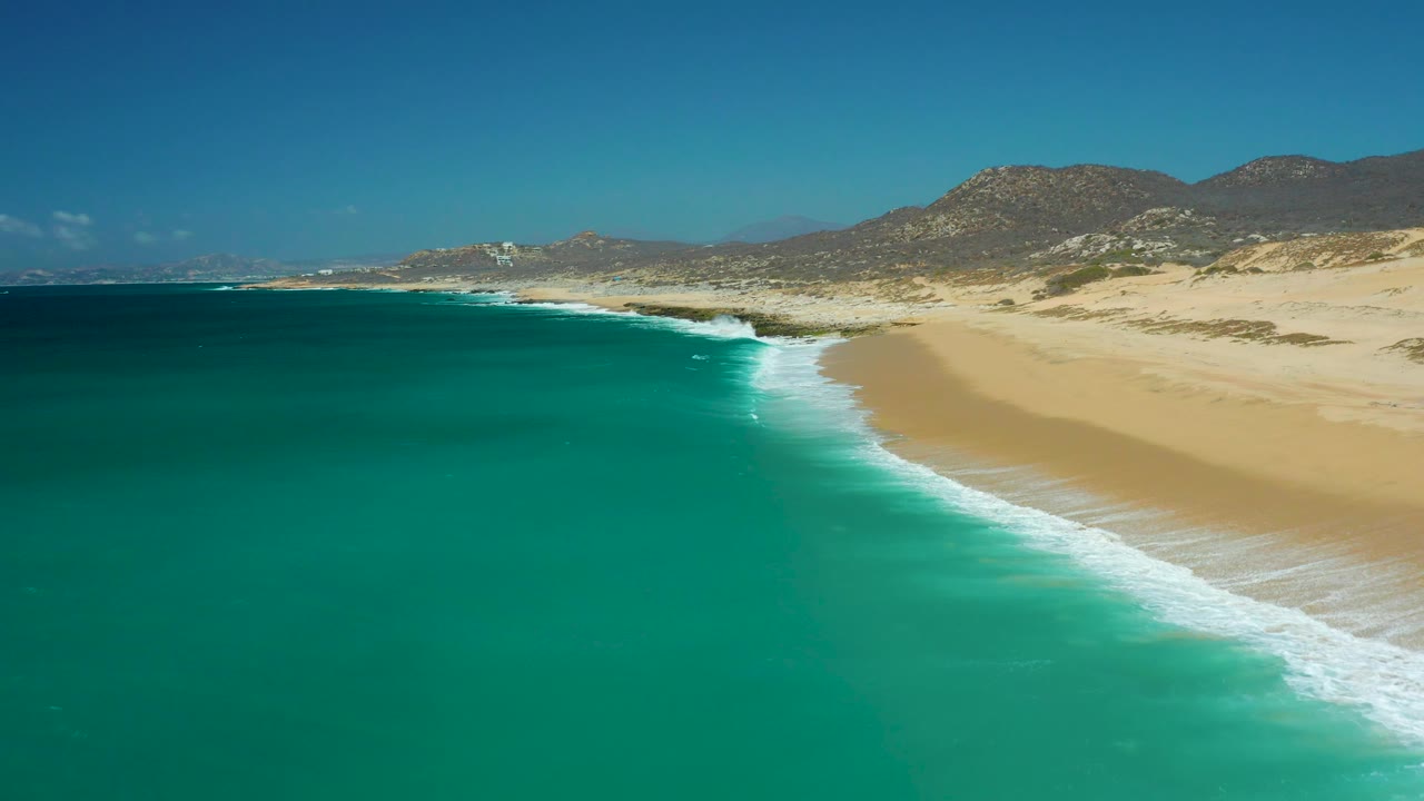 Aerial view along La Laguna Beach in Los Cabos with darker turquoise water at left, white foam tracing the shore, open sand at right, distant mountains, and no visible people.