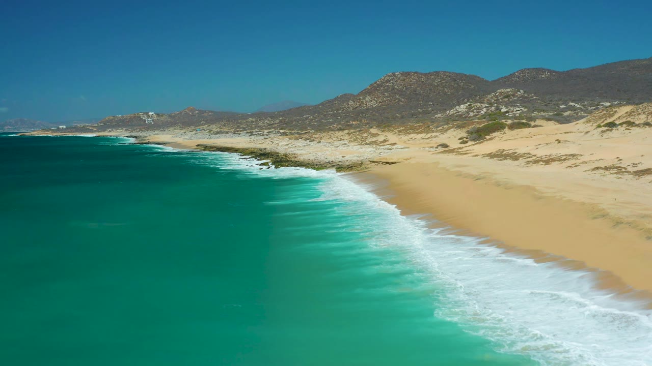 Aerial view one second in at La Laguna Beach Los Cabos, where the foamy shoreline curves across pale sand below dry rocky hills with no visible people.