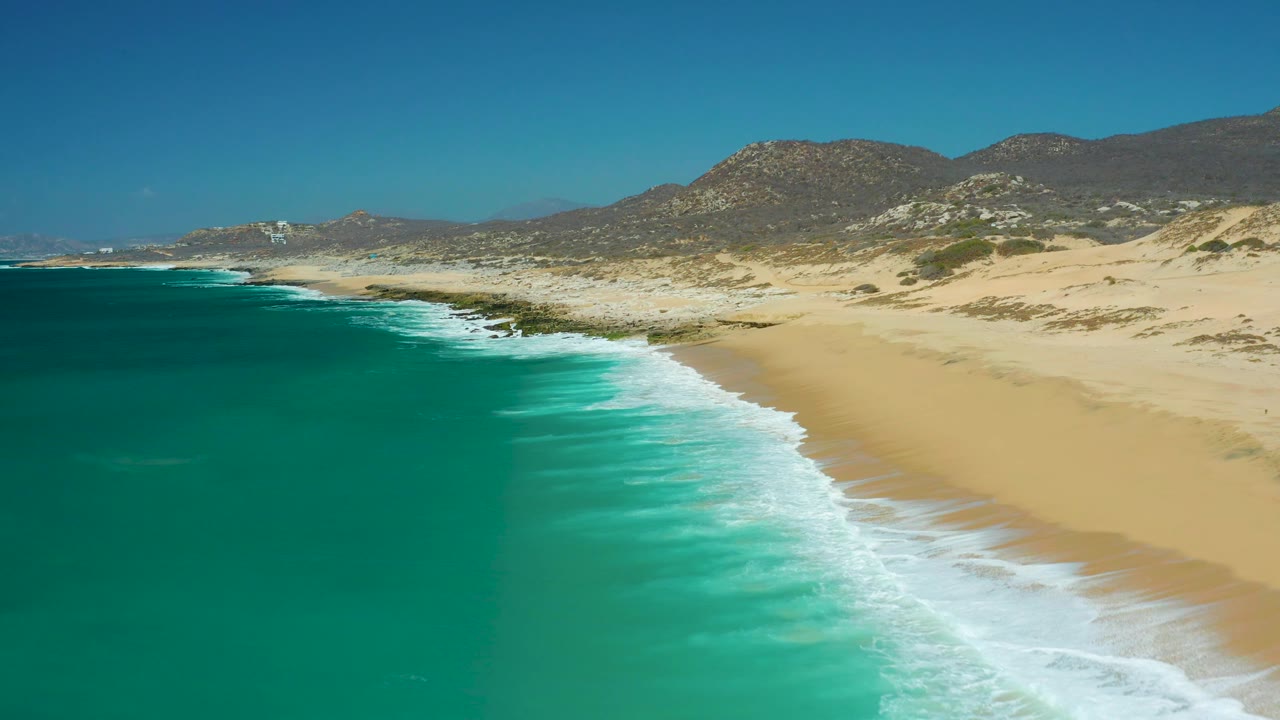 Aerial frame at 0:02 shows La Laguna Beach Los Cabos with more open water at left, a bright foam band along the shore, dry sand to the right, and no visible people.