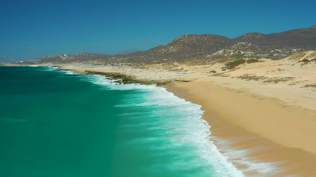 At 0:03, the La Laguna Beach Los Cabos shoreline fills the frame with turquoise water left, foam moving onto sand, dune patches inland, and no visible people.