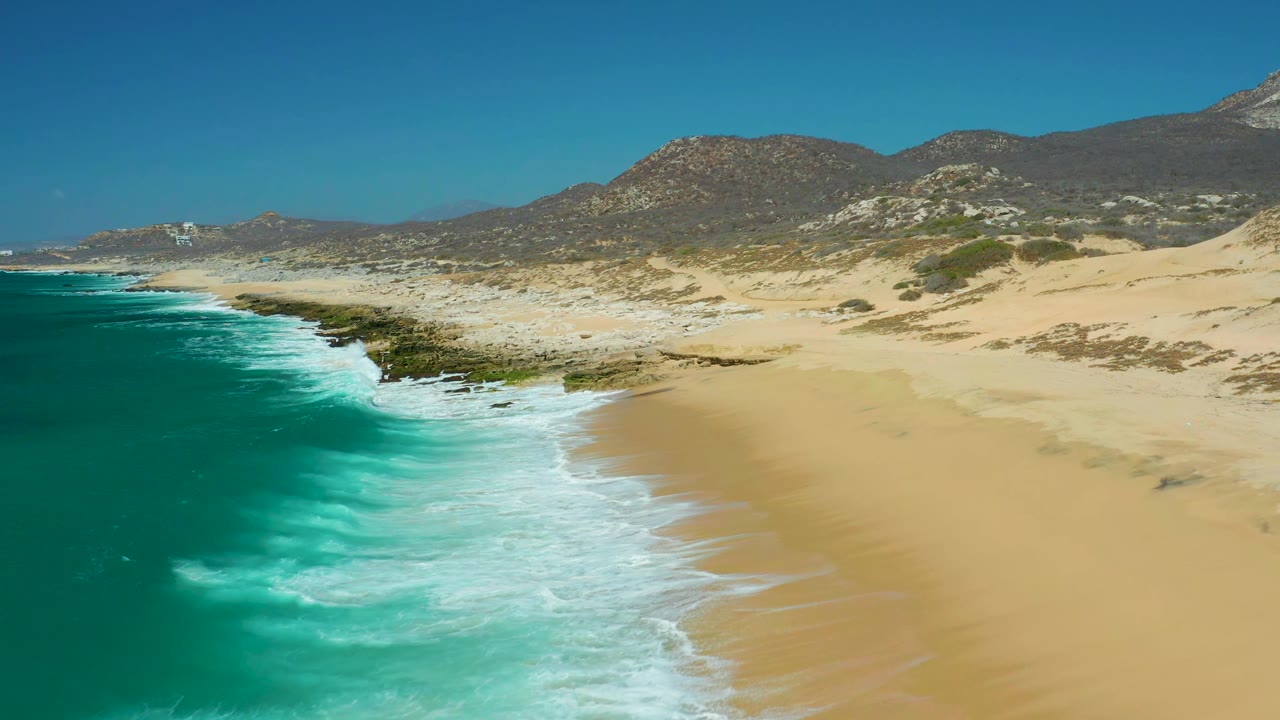 La Laguna Beach Los Cabos shoreline with foamy water pushing farther onto the sand, dark rocks tracing the coast, pale dunes behind the beach, distant mountains, clear sky, and no visible people.
