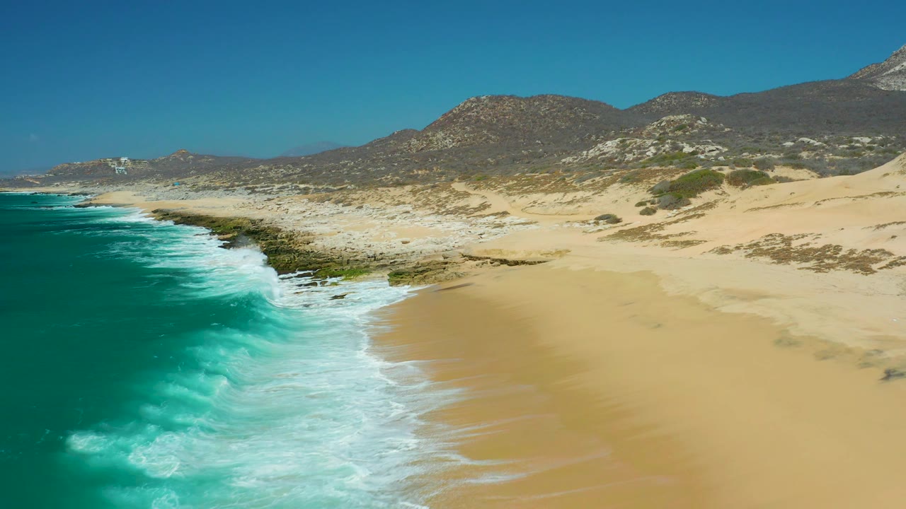 Aerial coastal frame at La Laguna Beach Los Cabos with a larger band of white foam curling toward the lower beach, turquoise water to the left, exposed rocks near center, dunes and mountains beyond, and no visible people.