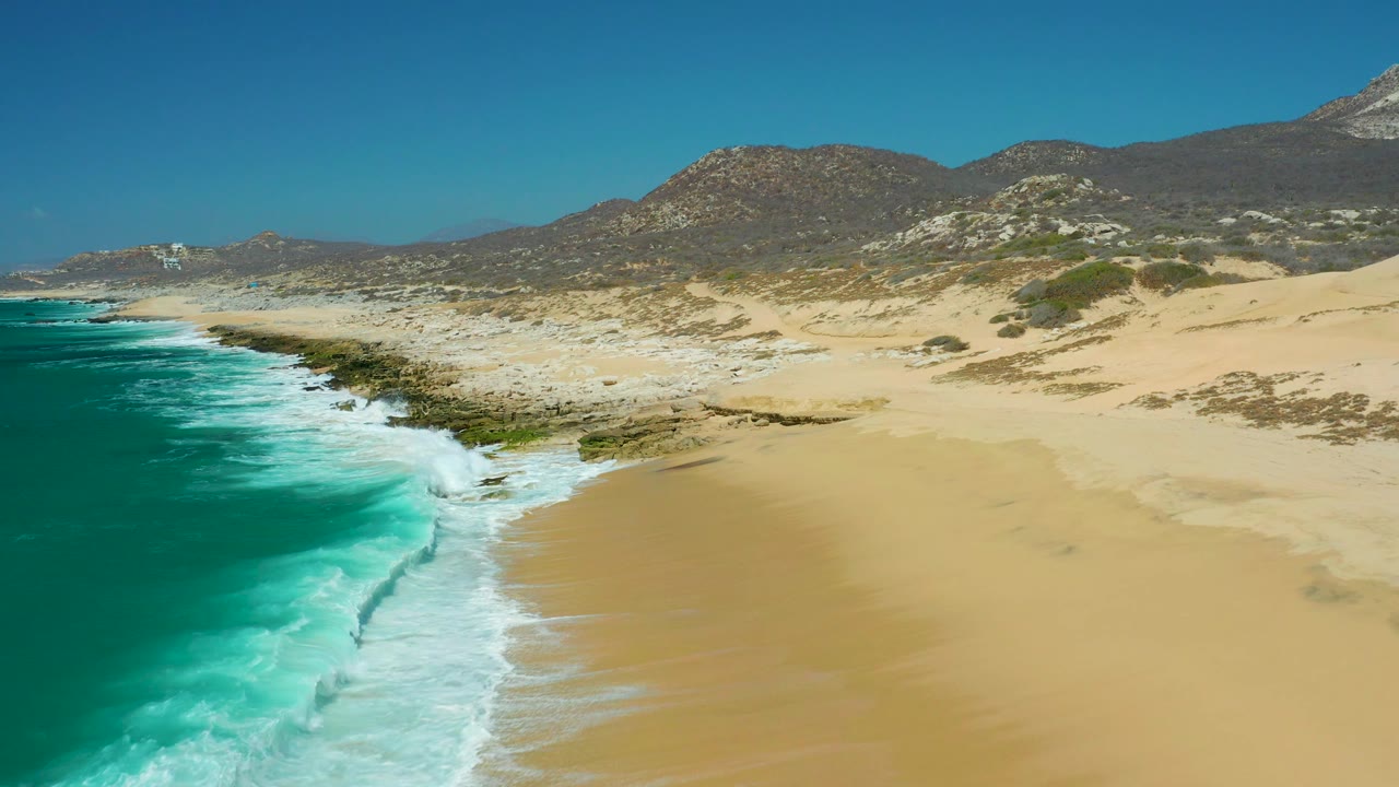 La Laguna Beach Los Cabos seen from above with the ocean filling the left edge, a fresh line of white foam at the shore, open sand to the right, rocky shelves ahead, scrub-covered dunes, mountains, and no visible people.