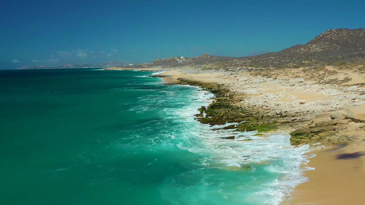 White foam spreads across the rocky shoreline at La Laguna Beach Los Cabos while the turquoise water and pale beach remain clearly visible, with no visible people.