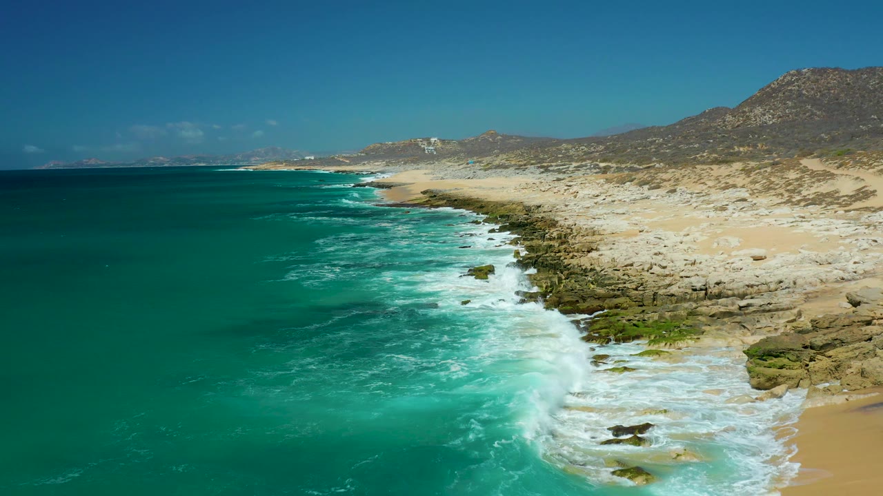 Foamy turquoise water washes along the rocky coastal edge at La Laguna Beach Los Cabos, with a pale sandy beach and low hills beyond, and no visible people.