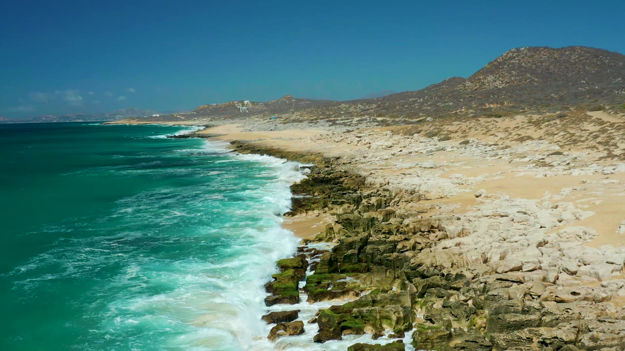 White waves push farther along dark rocks beside a sandy beach.