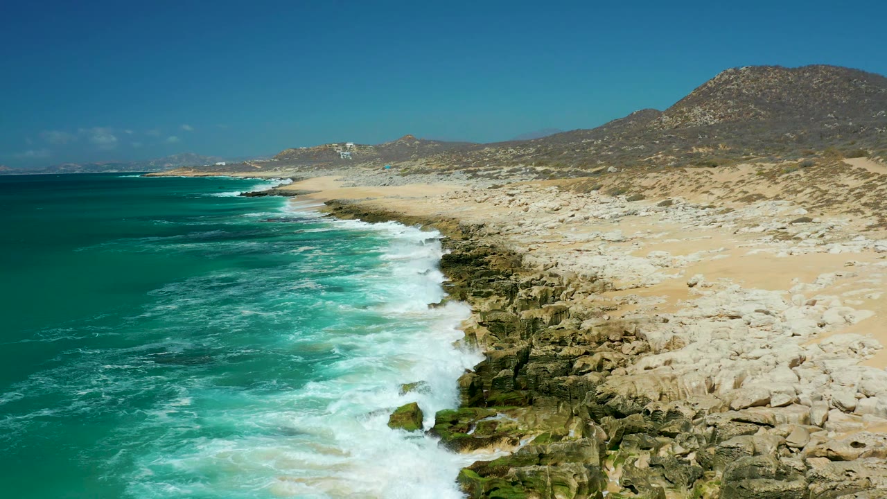 La Laguna Beach Los Cabos coast with foamy surf wrapping around foreground rocks, open turquoise water to the left, sandy beach and scrubby hills to the right, and no visible people.