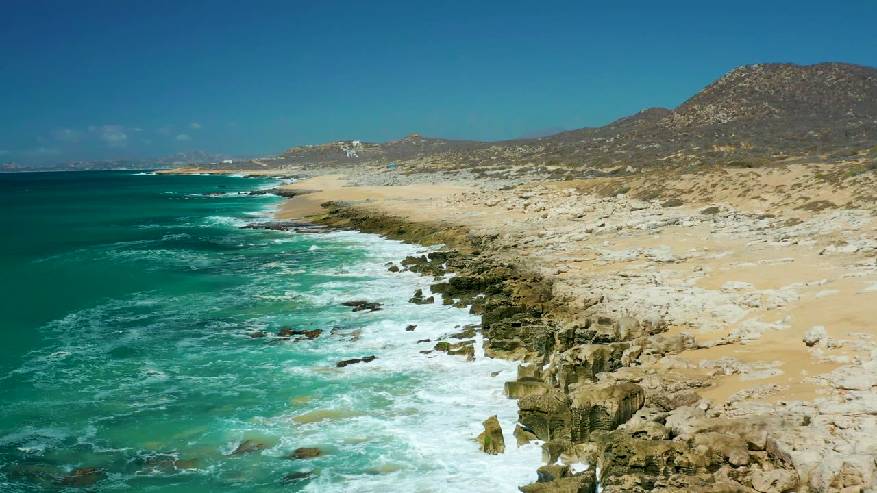 White foam spreads across shoreline rocks at La Laguna Beach Los Cabos while turquoise water, pale sand, hills, and clear sky remain visible with no visible people.