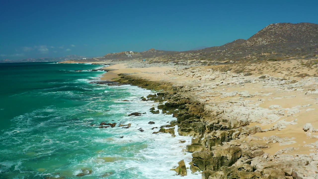 A fresh line of surf curls along the rocky La Laguna Beach Los Cabos shoreline, with teal water left of frame, sand and hills to the right, and no visible people.