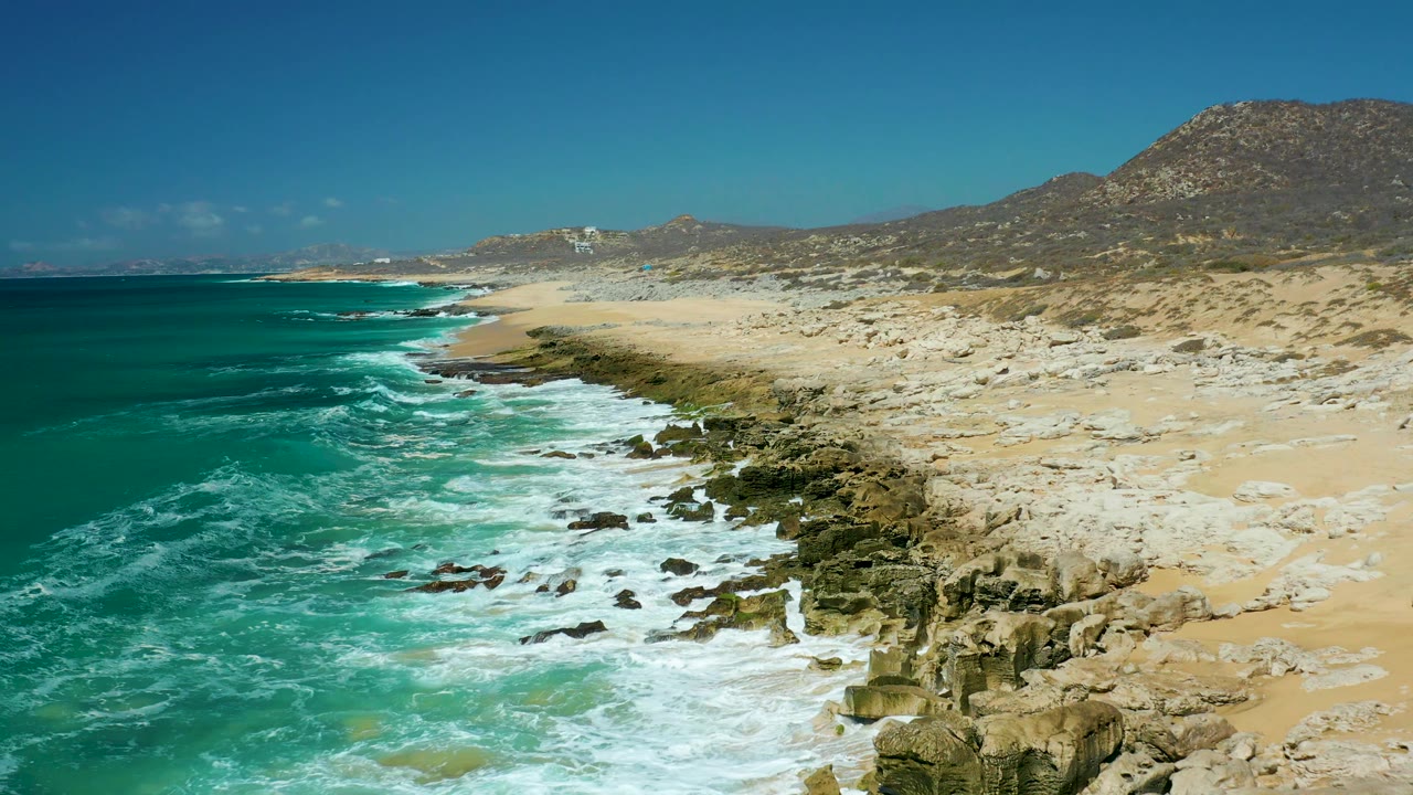 The elevated view looks down the La Laguna Beach Los Cabos coast as white surf washes over dark rocks beside an empty sandy beach and arid hills.