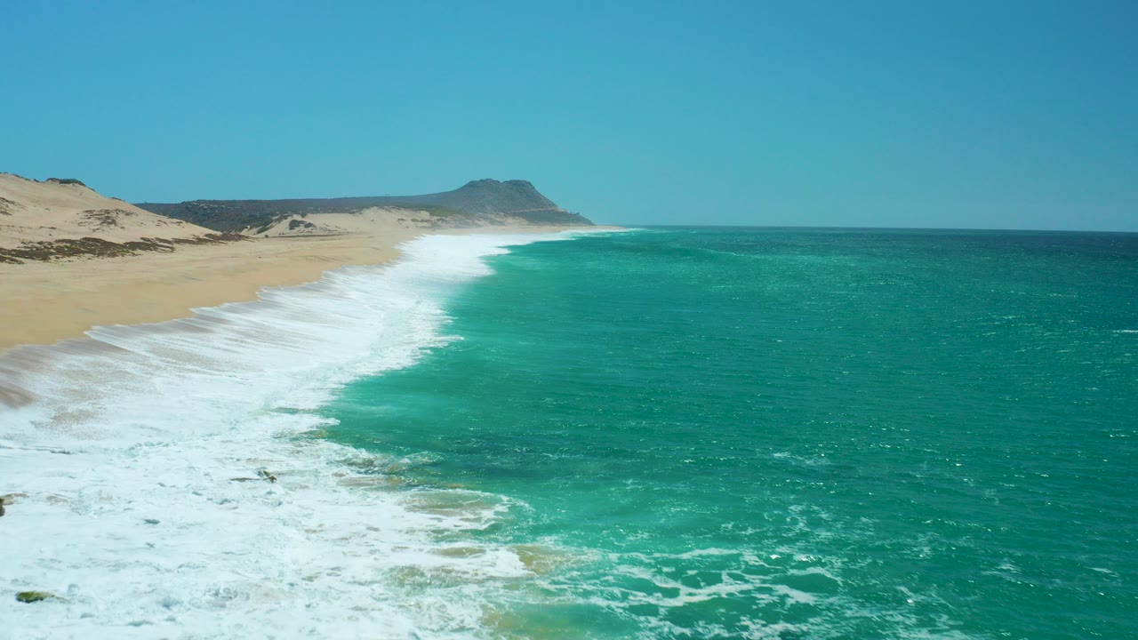 Aerial view of turquoise surf meeting a sandy beach under clear sky.