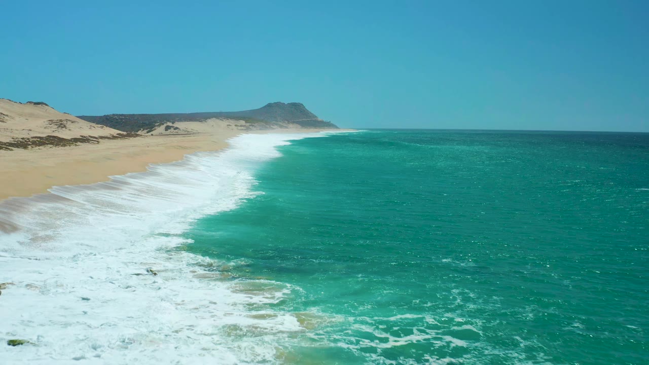 At 0:01, La Laguna Beach surf spreads wider across the sand while the turquoise ocean fills the right half of the aerial view, with dunes and a low headland beyond and no visible people.