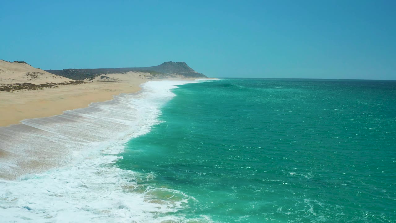 At 0:02, the foamy break forms a curved band along La Laguna Beach, with open turquoise water to the right, pale sand and dunes to the left, distant headland, and no visible people.