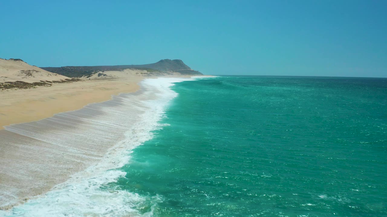 At 0:03, the aerial view looks down the La Laguna Beach coastline as thinner foam trails over wet sand, bright turquoise water sits at right, dunes line the left edge, and no visible people appear.
