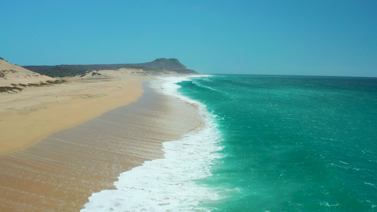 Aerial view of sandy beach, turquoise ocean, white surf, dunes, and clear sky.