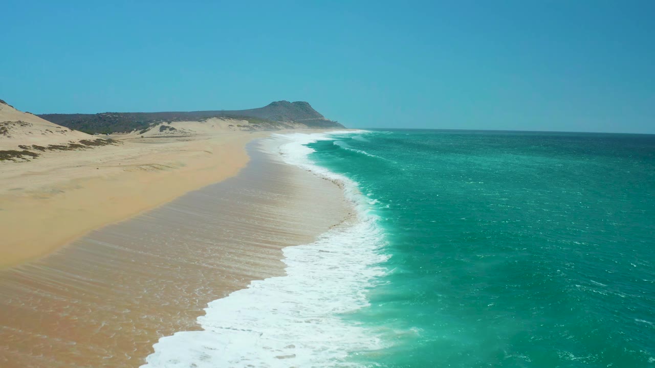 Wide aerial frame at La Laguna Beach with the surf band slightly farther along the sand, turquoise ocean filling the right side, dunes beyond the beach, clear sky, and no visible people.