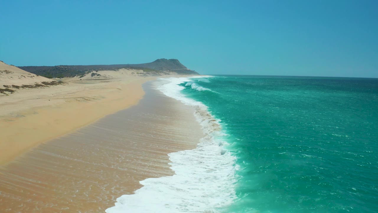 At La Laguna Beach, a stronger white wave breaks near the center-right of the frame, with open sand to the left, turquoise ocean to the right, distant dunes, and no visible people.