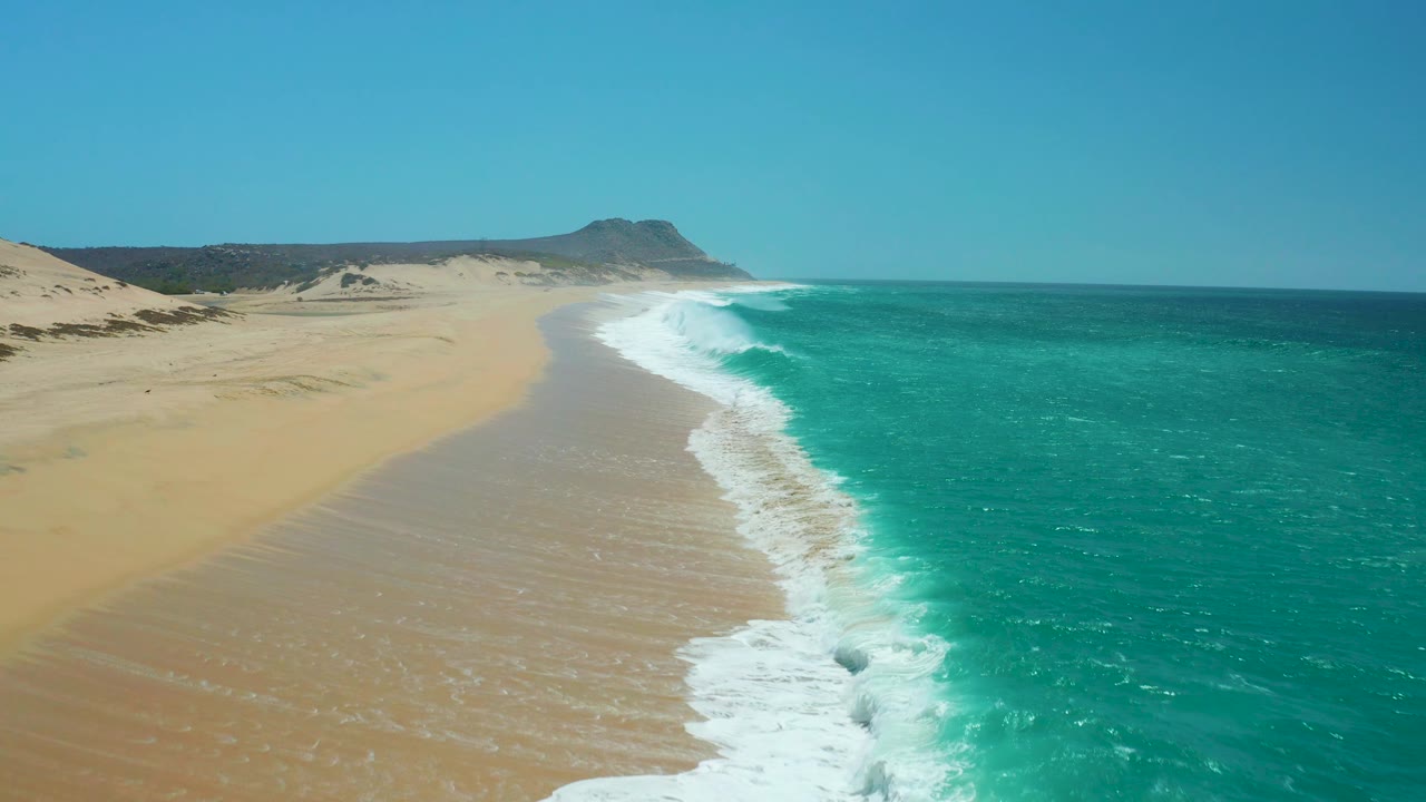 Aerial view over La Laguna Beach with wet sand forming a smooth curve under the surf, blue-green ocean stretching to the right, dunes in the background, and no visible people.