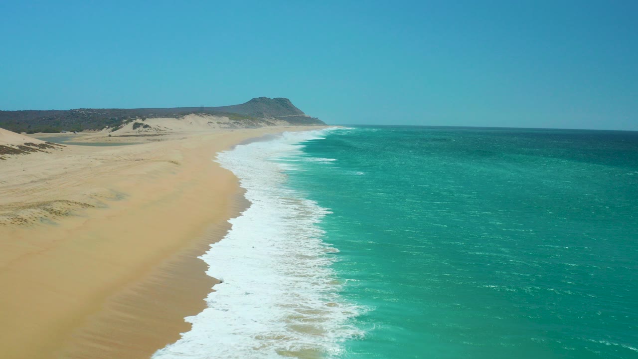 Aerial view at La Laguna Beach Los Cabos with golden sand on the left, turquoise water on the right, white surf curving toward a distant headland, and no visible people.