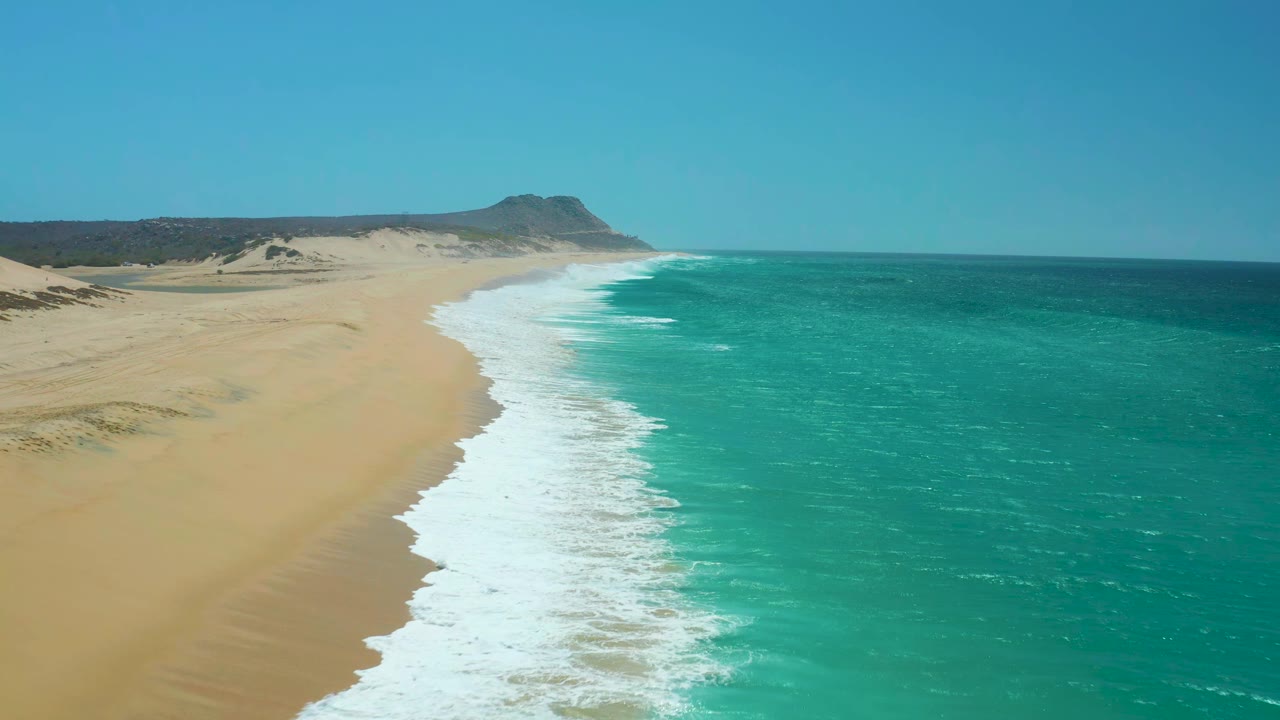 Drone view at La Laguna Beach Los Cabos where the foamy wave line sits slightly farther down the beach, with dunes behind the sand and no visible people.