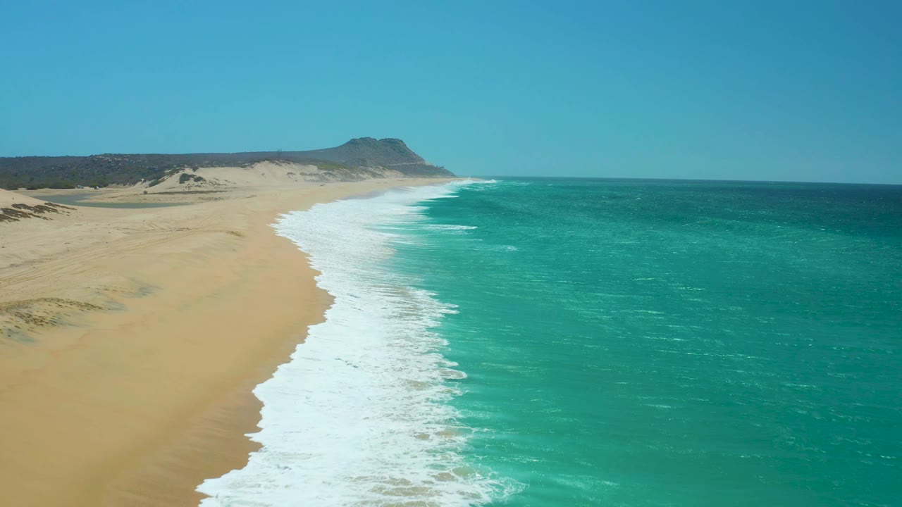Aerial coastline at La Laguna Beach Los Cabos with a broader band of white foam in the center, open sand to the left, clear blue water to the right, and no visible people.