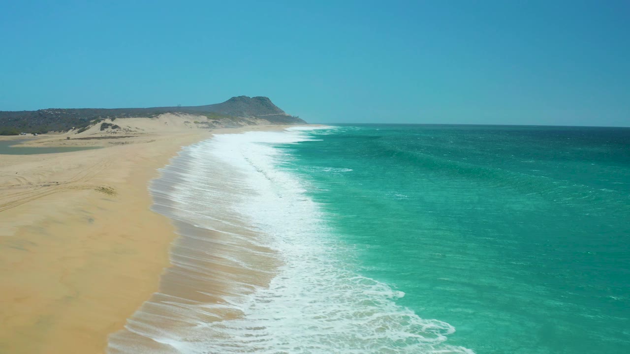 Aerial frame at La Laguna Beach with a curved band of breaking surf, pale sand on the left, turquoise ocean on the right, and no visible people.