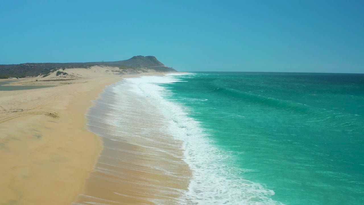 Aerial view of La Laguna Beach with more exposed sand in the foreground, foam trailing along the shoreline, ocean to the right, and no visible people.