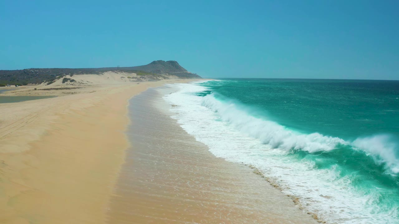 Aerial frame over La Laguna Beach, Los Cabos, Mexico, with a curving band of white foam between tan sand and turquoise water, no visible people.