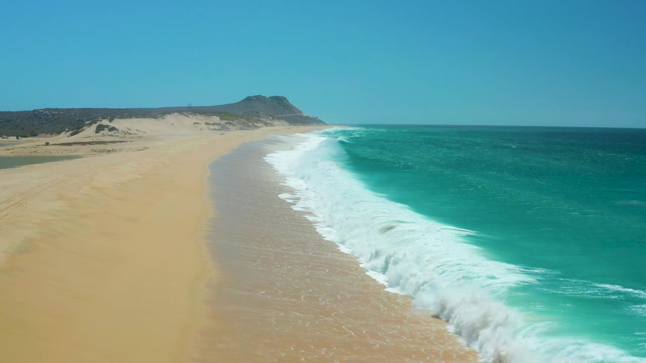 La Laguna Beach, Los Cabos, Mexico, seen from above as white surf washes up the long tan shoreline, with open water to the right and no visible people.
