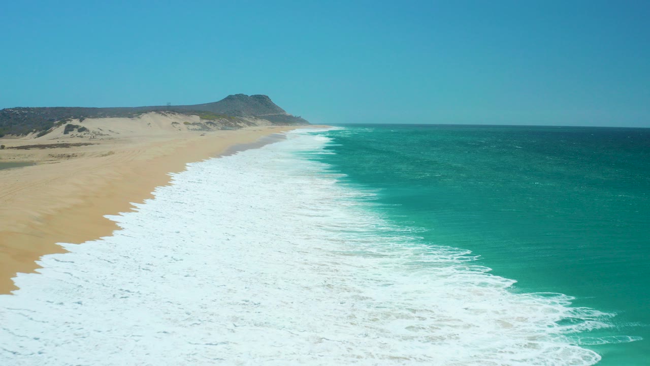 Aerial view of La Laguna Beach in Los Cabos with white surf spreading along sand, turquoise ocean to the right, a distant hill on the horizon, and no visible people.