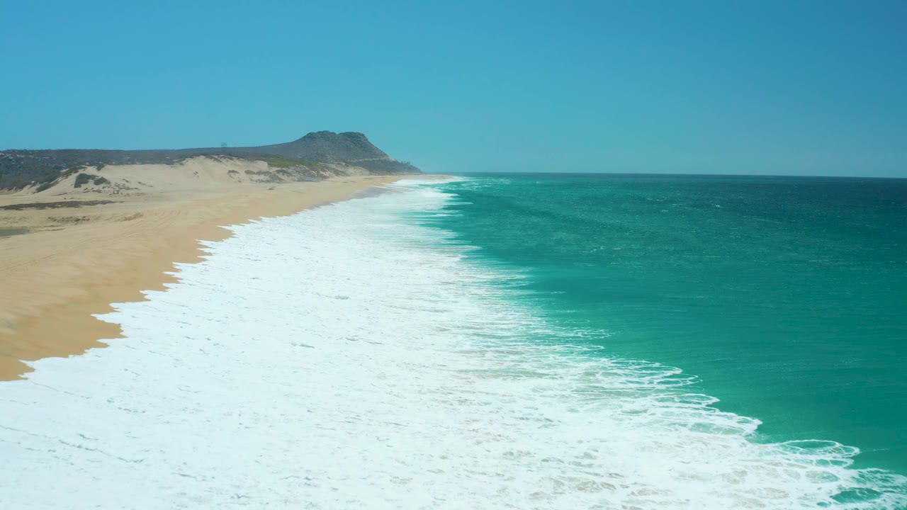 High aerial frame of La Laguna Beach with a long band of white breaking waves between tan sand and turquoise sea, a low hill in the distance, and no visible people.