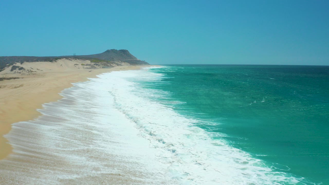 Aerial view of La Laguna Beach in Los Cabos with turquoise water breaking into white surf beside a broad sandy shore and low dunes, no visible people.