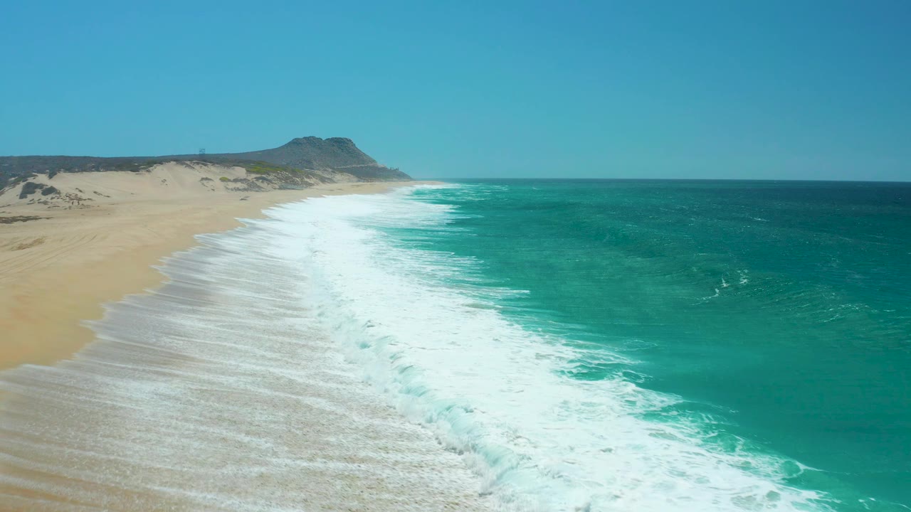 Elevated view of the La Laguna Beach coast with a fresh white wave band breaking diagonally along the sand and open teal water to the right, no visible people.