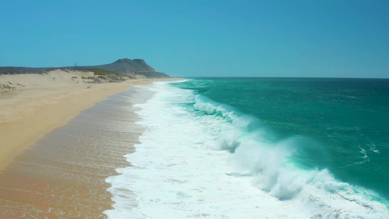 La Laguna Beach Los Cabos shoreline viewed from above as a fresh band of white foam rolls diagonally across the sand, with open turquoise water and no visible people.
