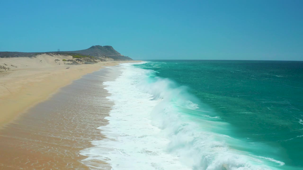 A larger white wave spreads across the La Laguna Beach shoreline, with bright turquoise water to the right, pale sand to the left, dunes in back, and no visible people.