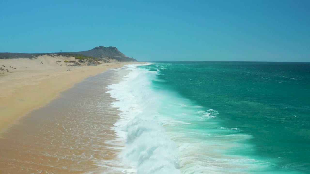 Oblique aerial view of La Laguna Beach Los Cabos as white surf washes along the empty sandy edge, with deeper teal water offshore and no visible people.
