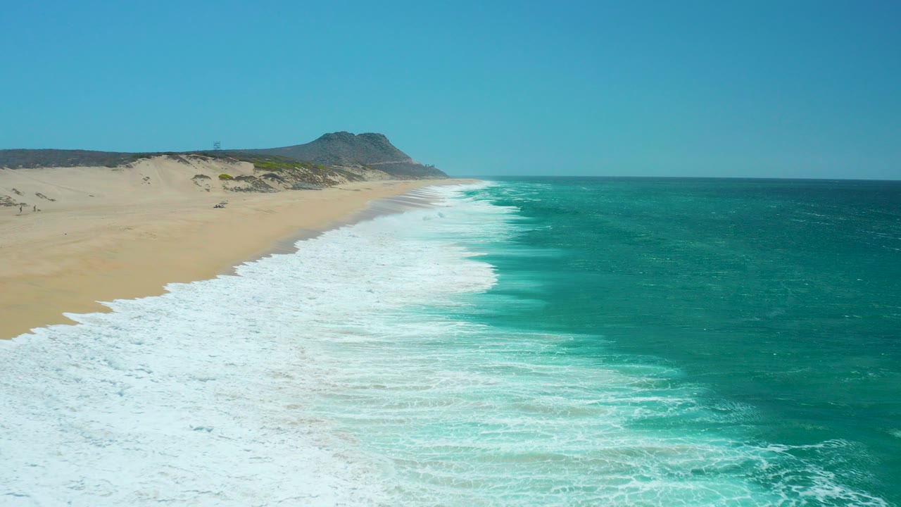 At La Laguna Beach in Los Cabos, Mexico, white surf spreads along the sandy shoreline with turquoise water to the right, a distant hill on the horizon, and no visible people.