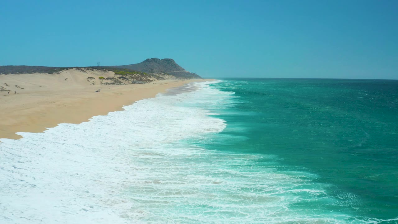 At La Laguna Beach in Los Cabos, Mexico, a wider sheet of white foam reaches up the beach, the ocean remains vivid turquoise to the right, and no visible people appear on the sand.