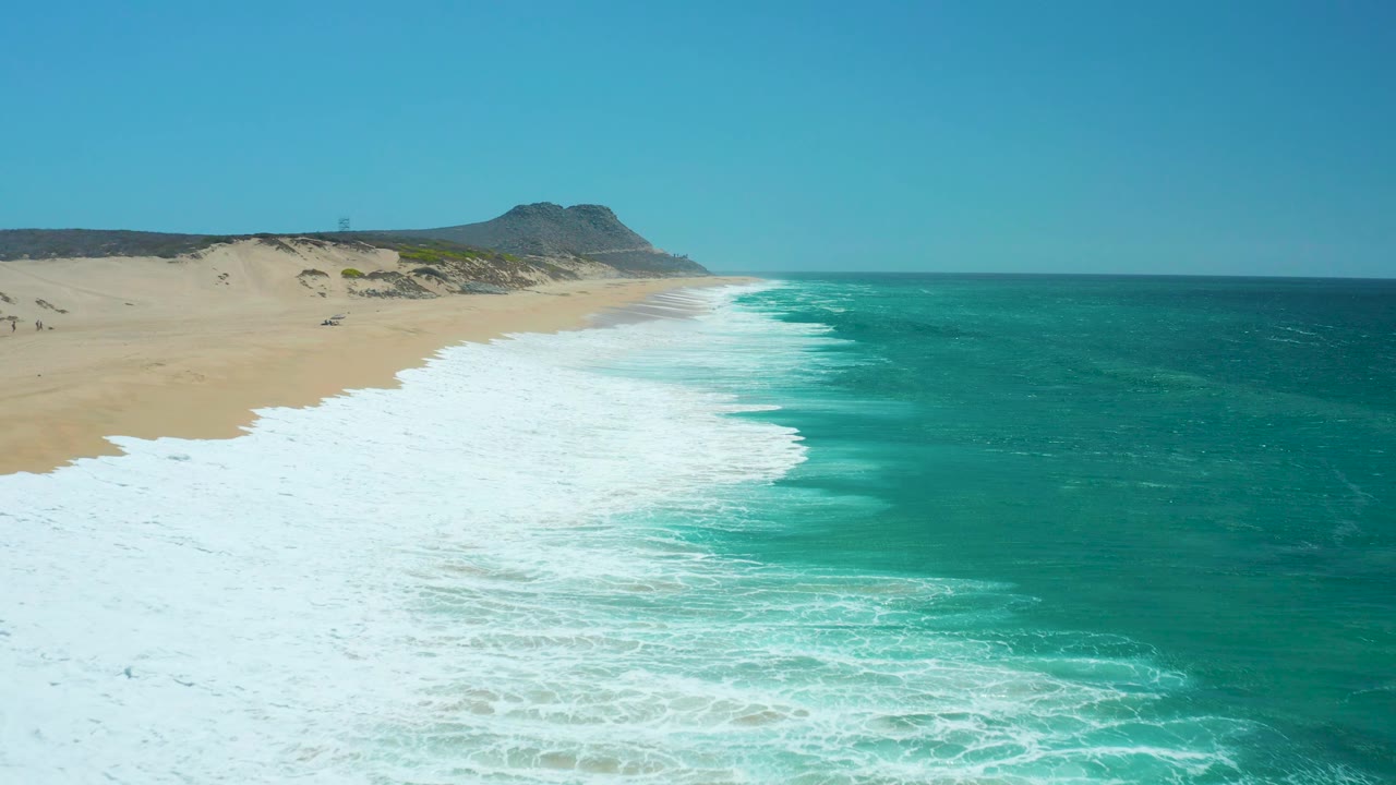 Aerial beach view with surf retreating and a distant hill beyond the sand.