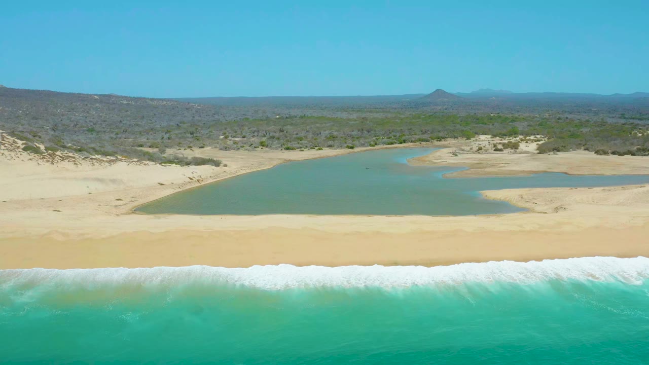 At 0:01, La Laguna Beach Los Cabos is seen from above with a broad band of turquoise water, fresh white foam along the beach, lagoon water set back from the sand, low dunes, vegetation, and no visible people.