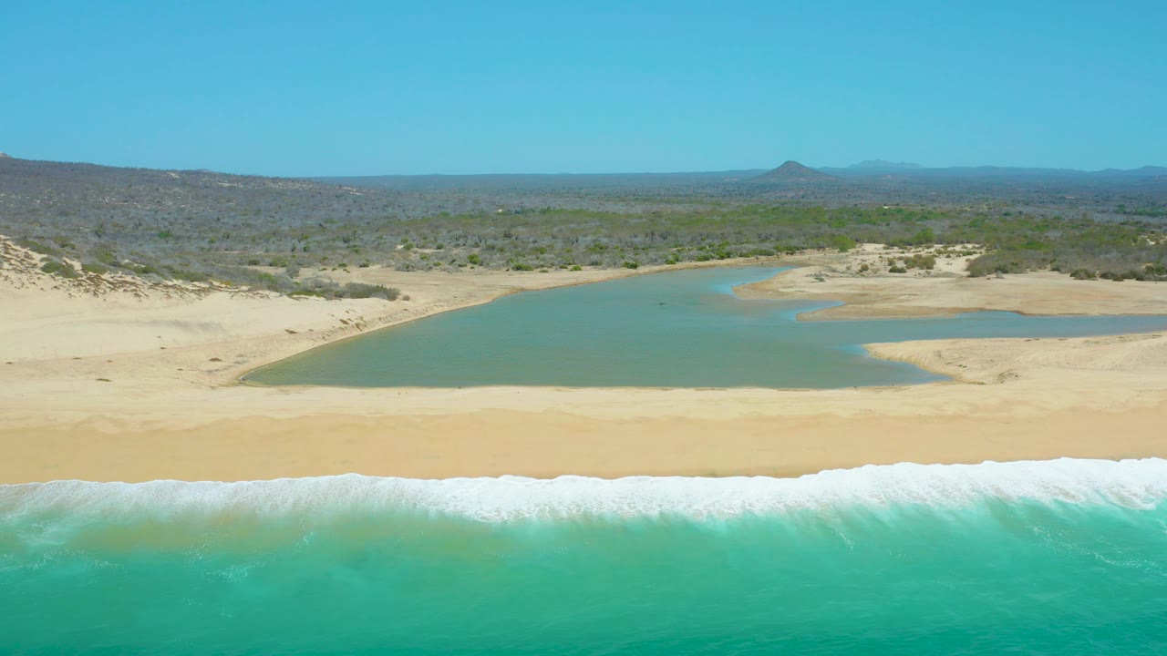 At 0:02, the aerial composition at La Laguna Beach Los Cabos keeps the ocean foreground, white foam band, sandy shoreline, calm lagoon, dune edge, scrubland, distant hills, and clear sky visible with no visible people.
