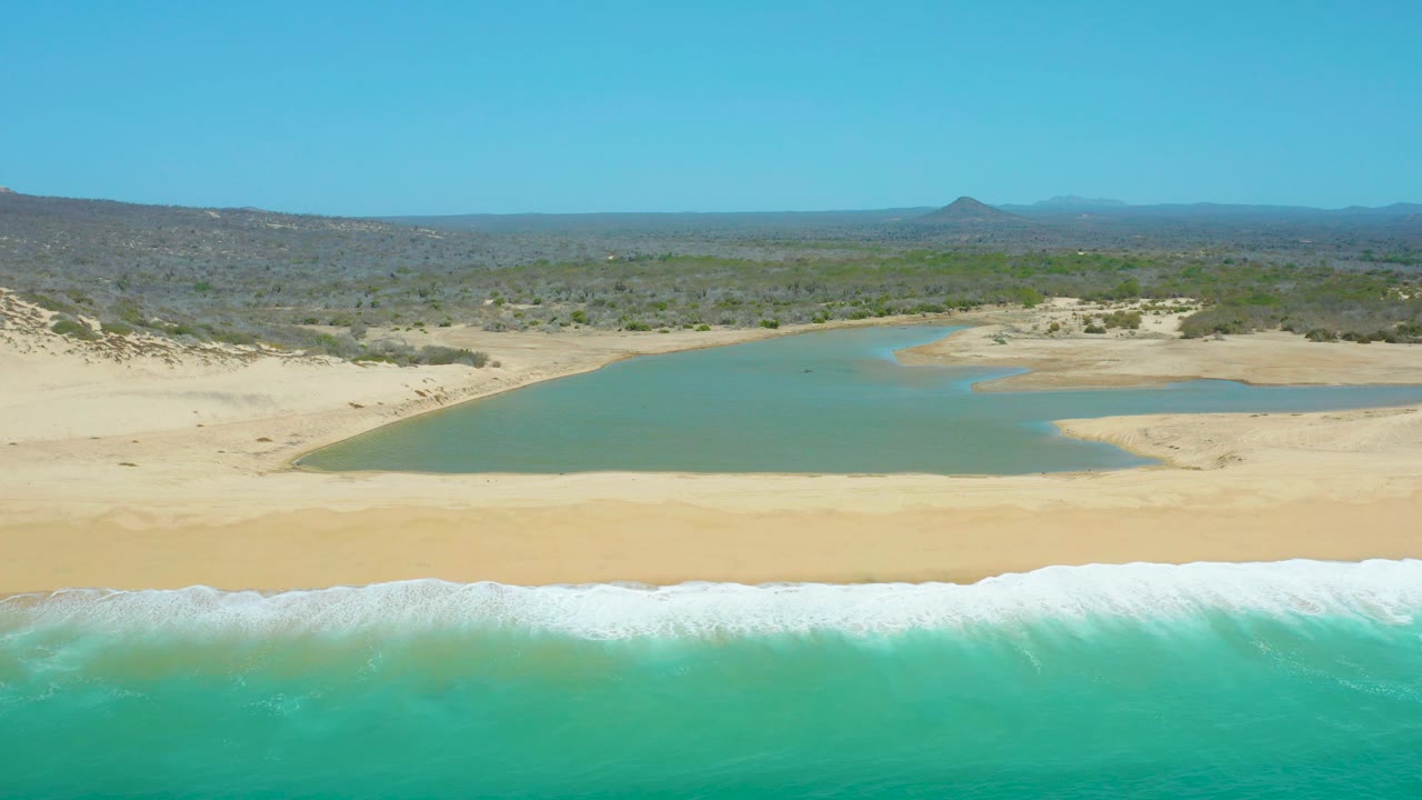 At 0:03, La Laguna Beach Los Cabos appears in a wide elevated frame with foamy turquoise surf at the bottom, pale beach, green-blue lagoon, wind-shaped dunes, low vegetation, distant hills, and no visible people.