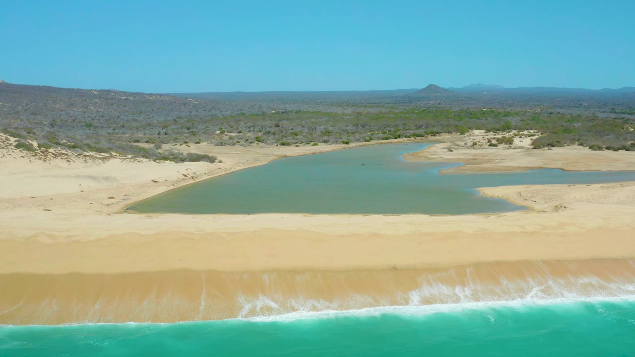 Aerial view at La Laguna Beach Los Cabos with the surf line lower in frame, a pale sandy beach, still lagoon water, dry scrubland, distant hills, clear sky, and no visible people.