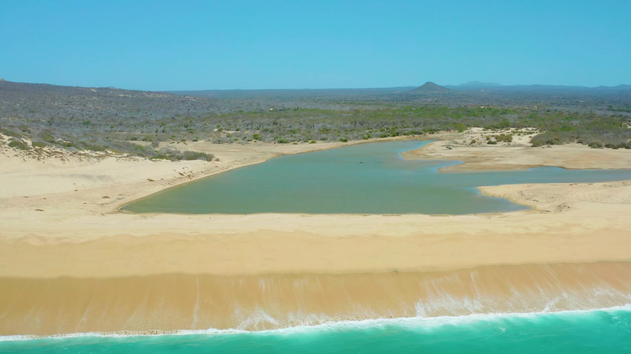 La Laguna Beach Los Cabos appears from above with turquoise surf crossing the lower edge, empty sand in the center, green lagoon water beyond, arid brushland, hills, and no visible people.
