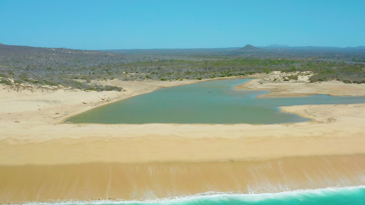 Aerial still from La Laguna Beach Los Cabos with ocean surf along the lower frame, a wide empty beach, a calm lagoon, low vegetation, hazy distant hills, blue sky, and no visible people.