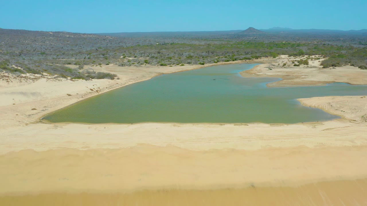 Aerial view at La Laguna Beach Los Cabos, Mexico, with the lagoon stretching across the frame, sandy banks in the foreground, scrubland behind it, distant hills, and no visible people.