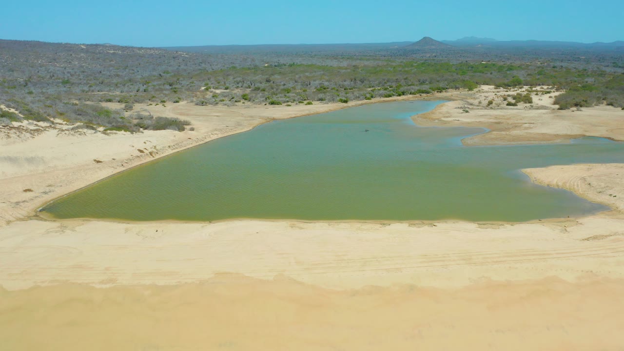 Mid-clip aerial view over La Laguna Beach Los Cabos, Mexico, with green lagoon water close to the foreground sand, scrub-covered terrain behind, distant hills, and no visible people.