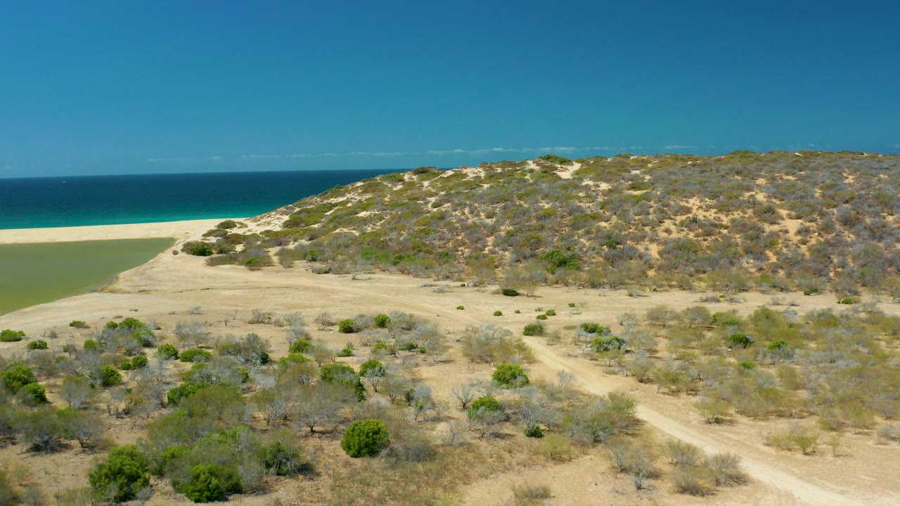 Opening aerial frame over La Laguna Beach Los Cabos, with dry scrub dunes in front, a pale sandy beach and green lagoon edge to the left, turquoise ocean beyond, and no visible people.