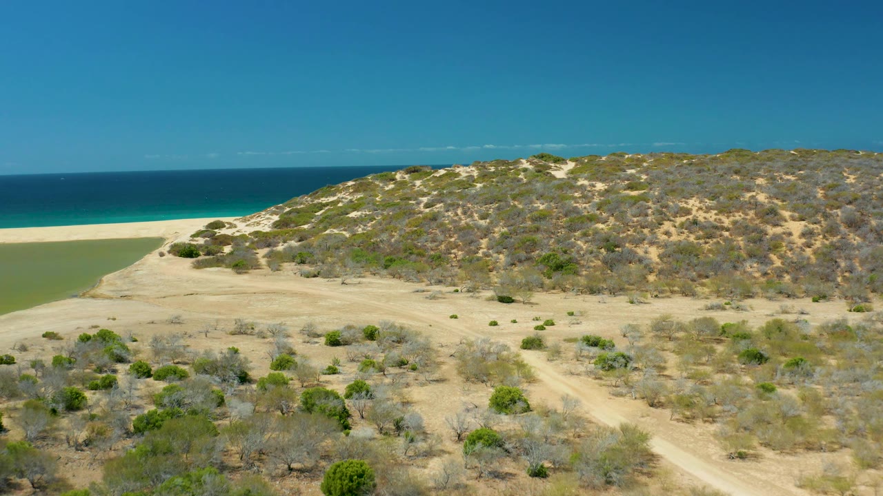 At 0:02, the La Laguna Beach Los Cabos aerial view places the scrub-covered dune ridge across the middle, with sandy flats below, lagoon color at left, ocean beyond, and no visible people.